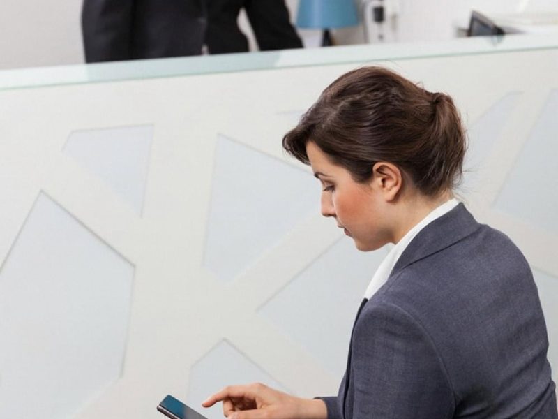 Traveler with red suitcase using her phone to check in at a hotel lobby while a receptionist works behind the desk