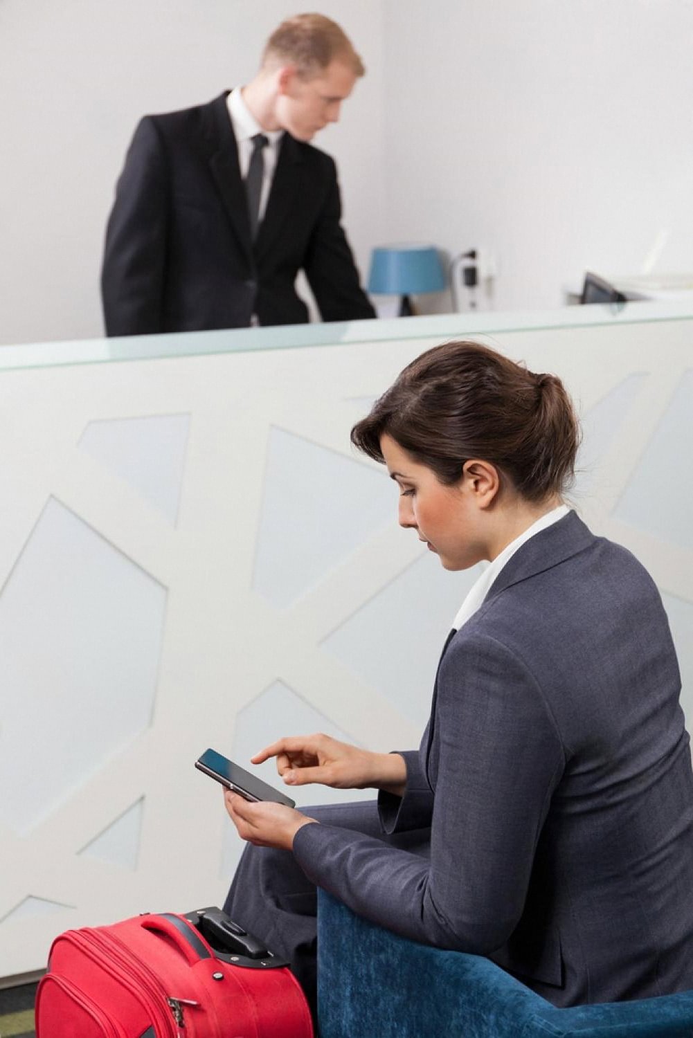 Traveler with red suitcase using her phone to check in at a hotel lobby while a receptionist works behind the desk